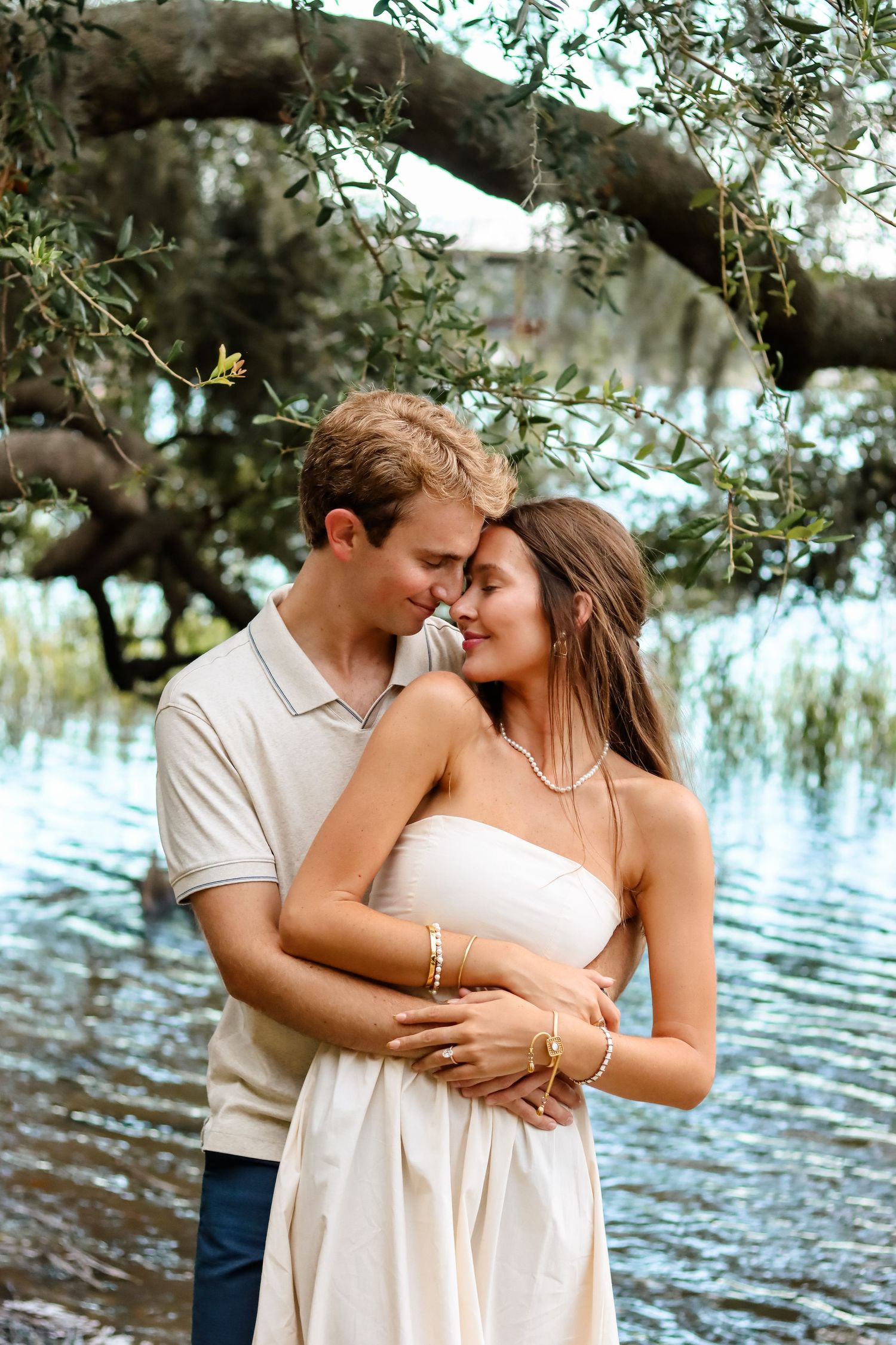 A romantic couple embraces under a willow tree by the waterfront.