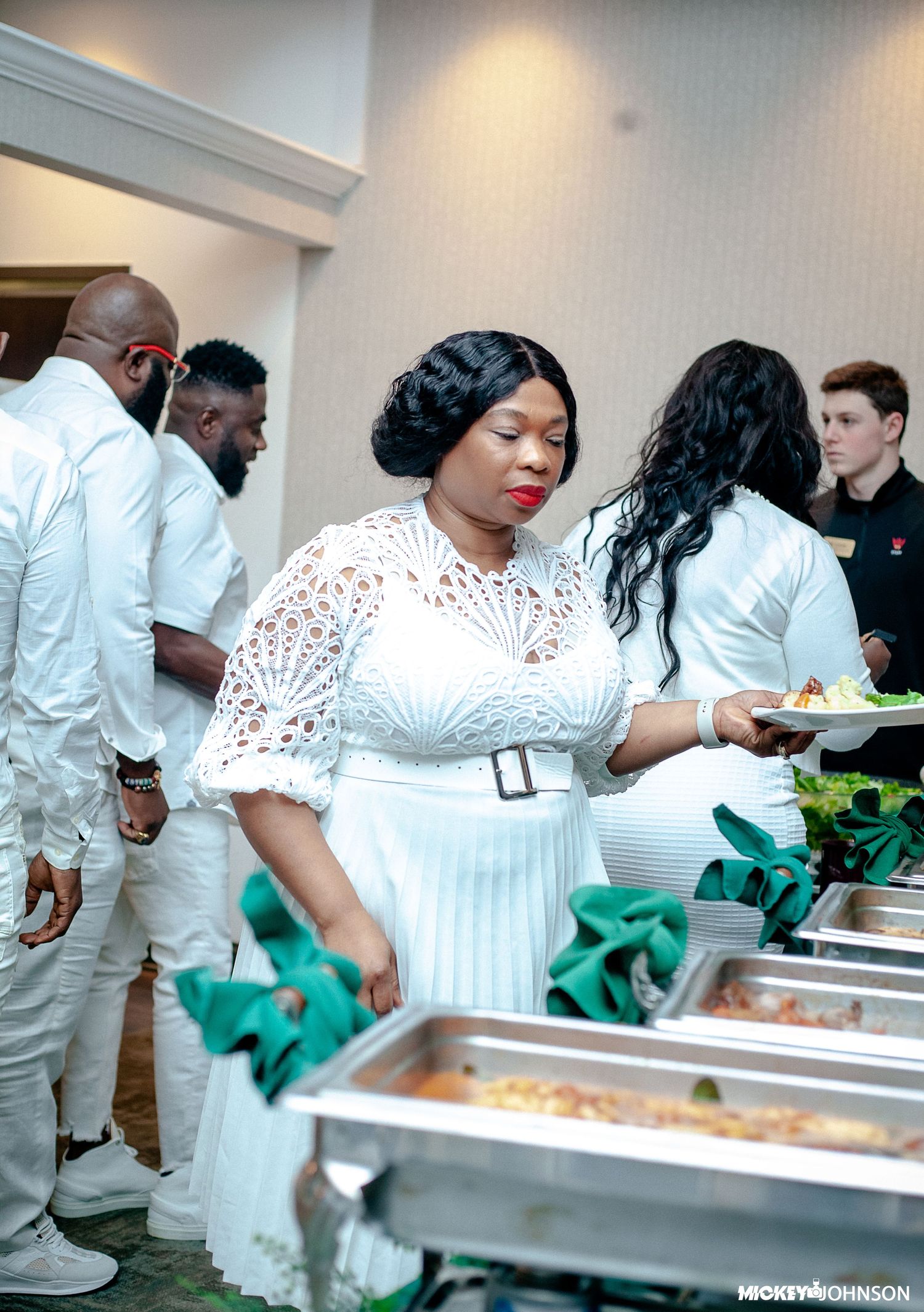 People in white attire serving food at buffet line with green accents.