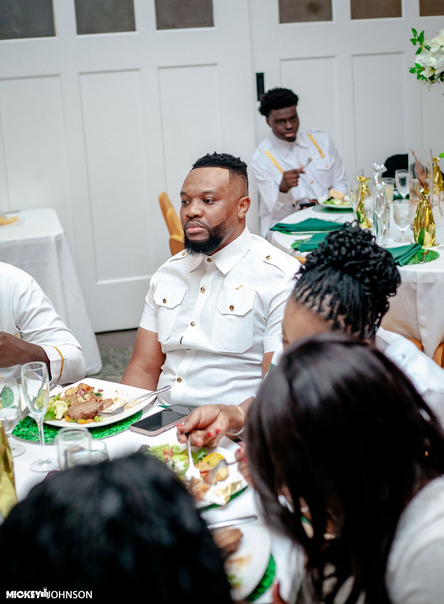 Guests in white attire seated at dining tables with green decorative elements.
