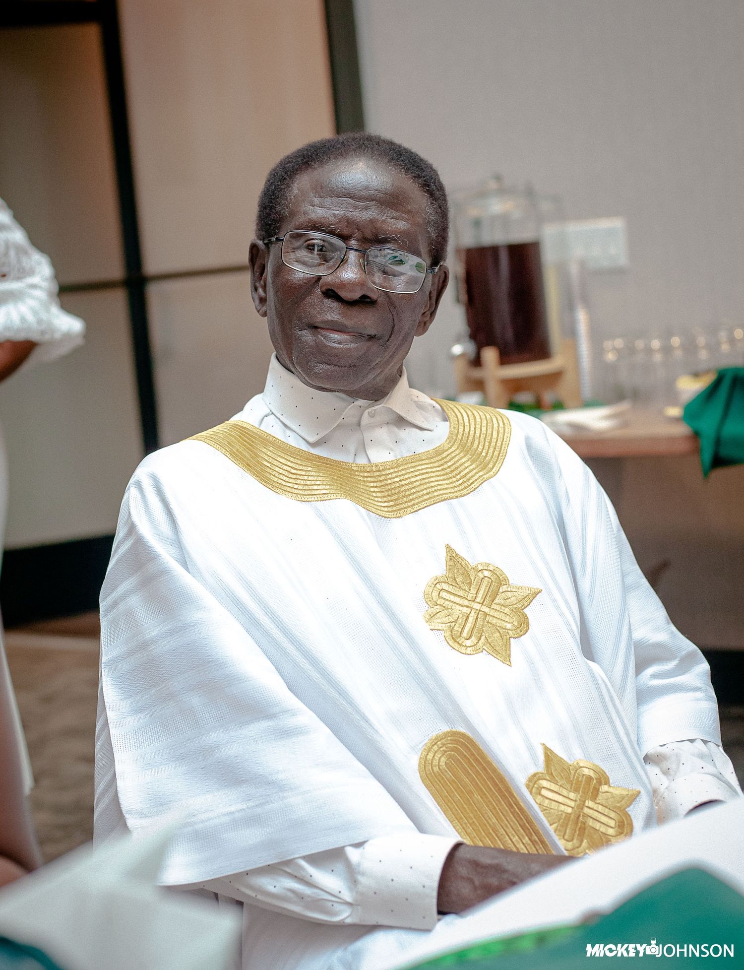 Person wearing traditional white and gold attire seated at formal event.