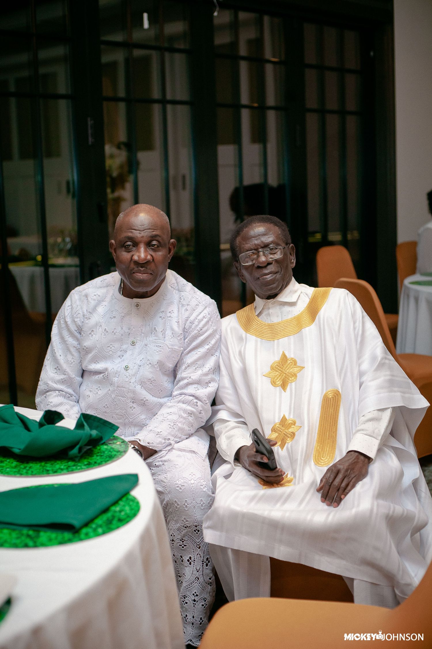 Two guests in traditional white and gold attire seated at a formal event.