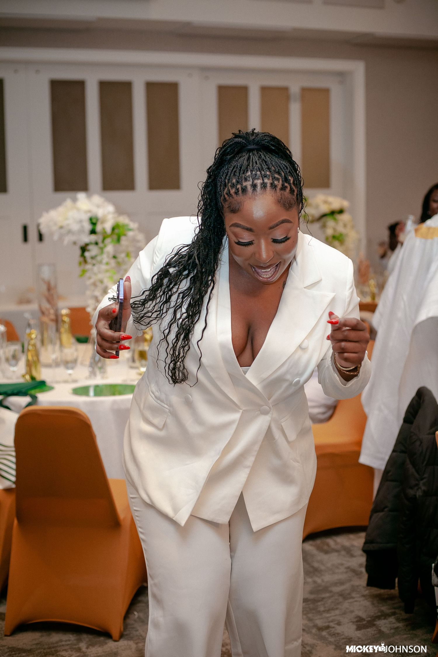 People in white attire dancing and celebrating at an indoor event with orange chairs.