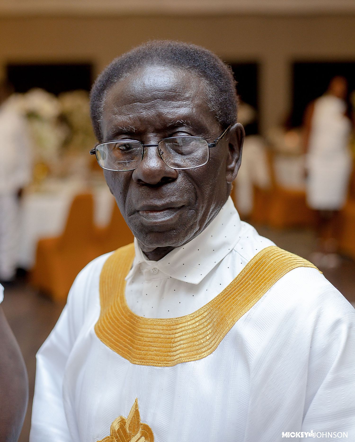 An elder wearing glasses and white attire with gold accents at a formal gathering.