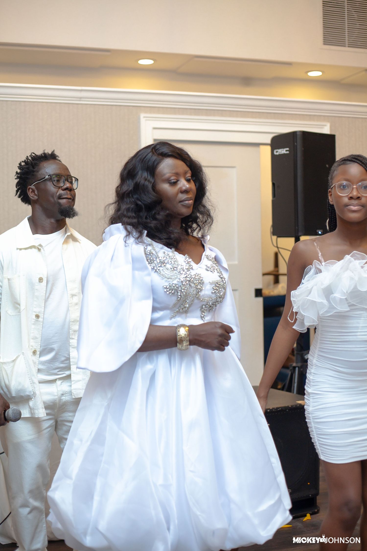 Three people in white outfits stand together at a formal event with stage lighting.