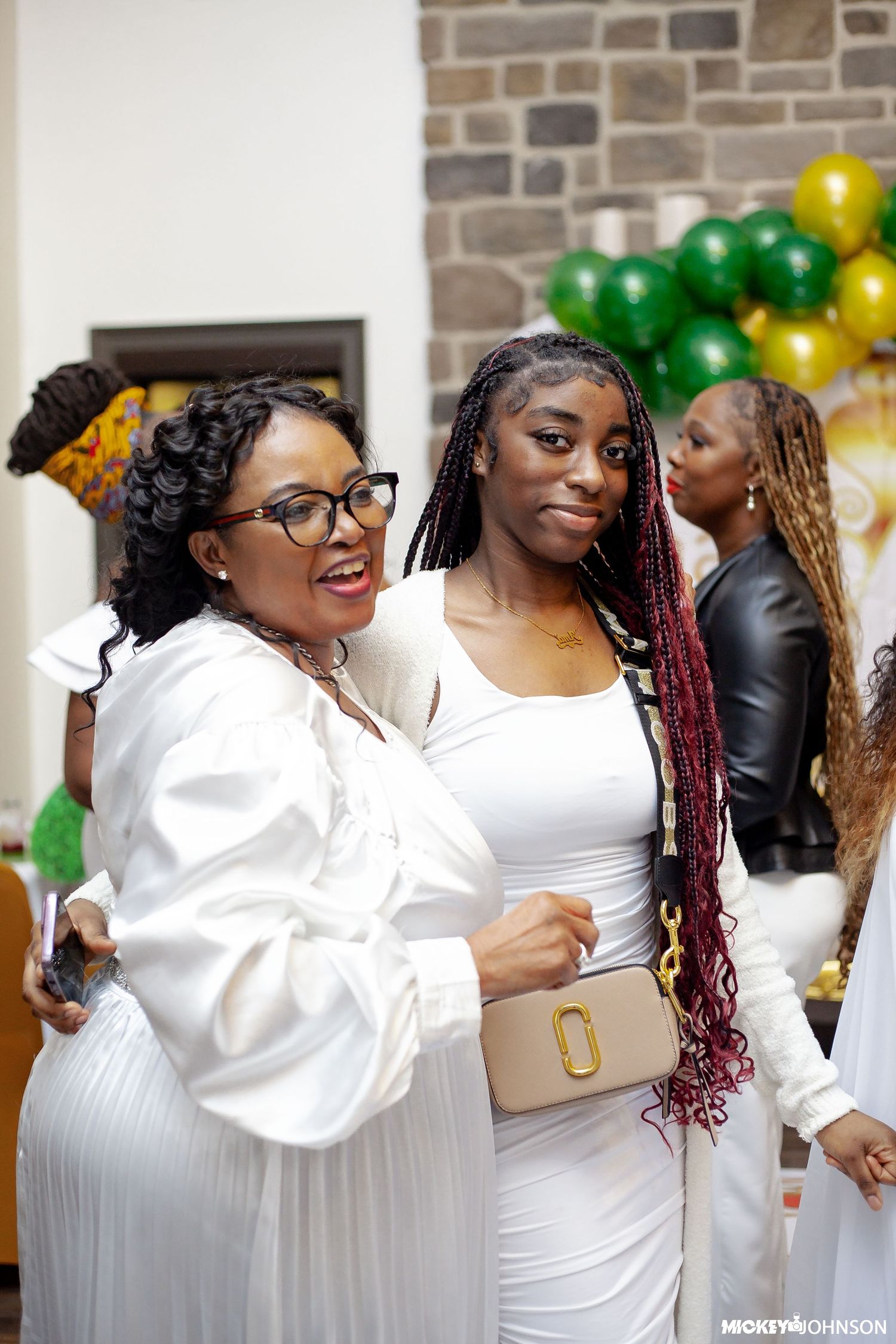 Guests in white attire celebrating near a brick wall with green and gold balloons.
