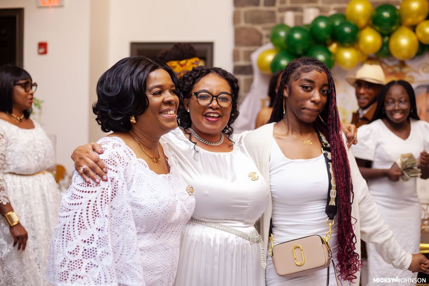 A group of people wearing white outfits smile and embrace at a celebration with green and gold balloons.
