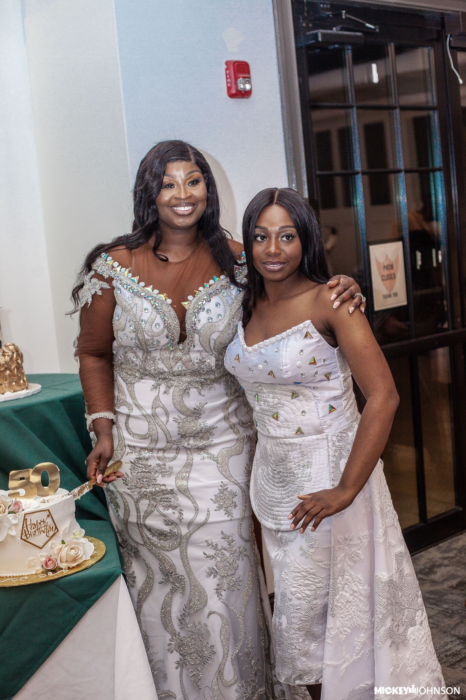 Two attendees in elegant white and silver formal dresses pose together by a green table with cake.