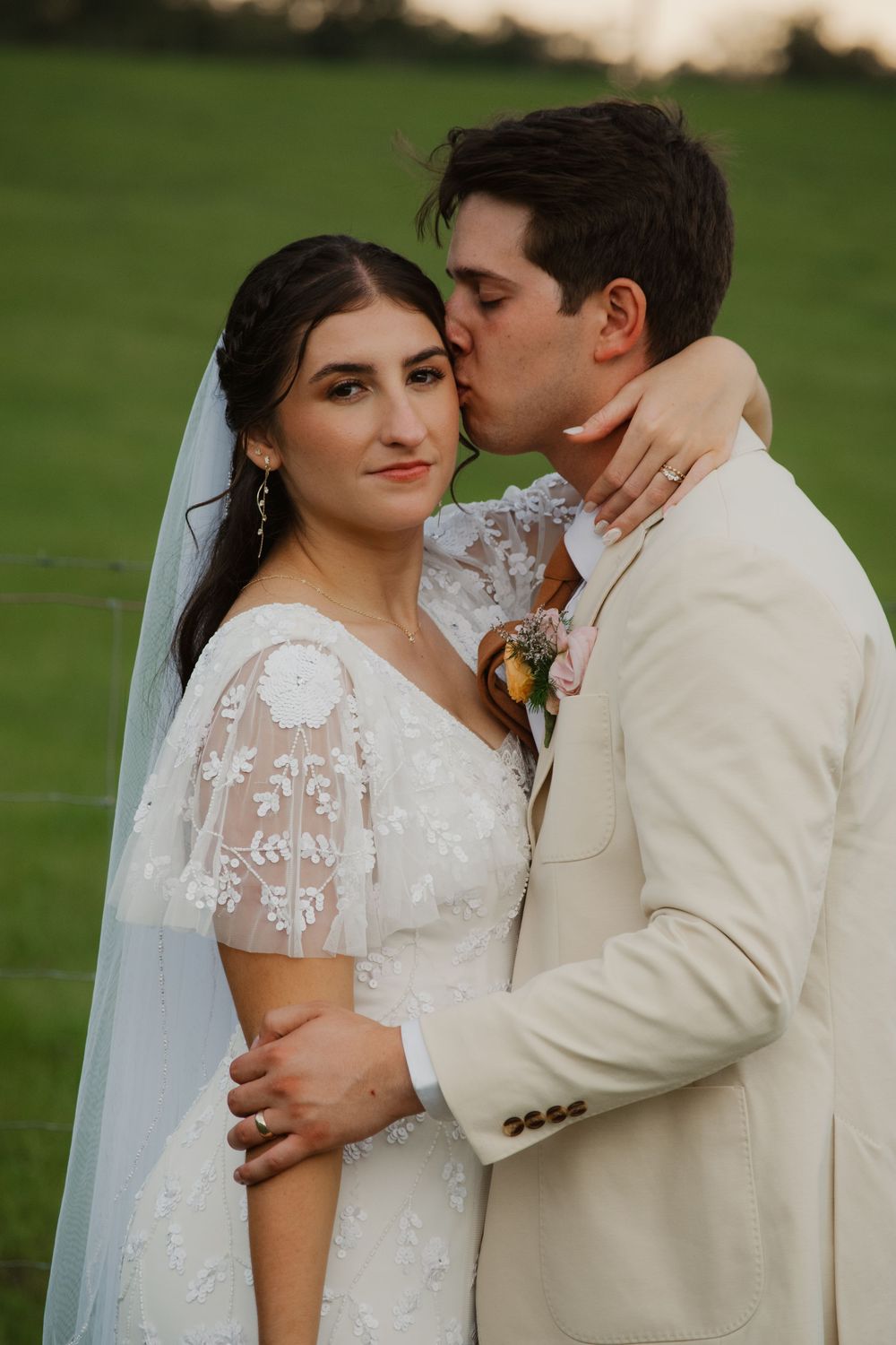 Groom in ivory suit tenderly kisses bride on cheek as she smiles at camera, standing in a lush green field at sunset.