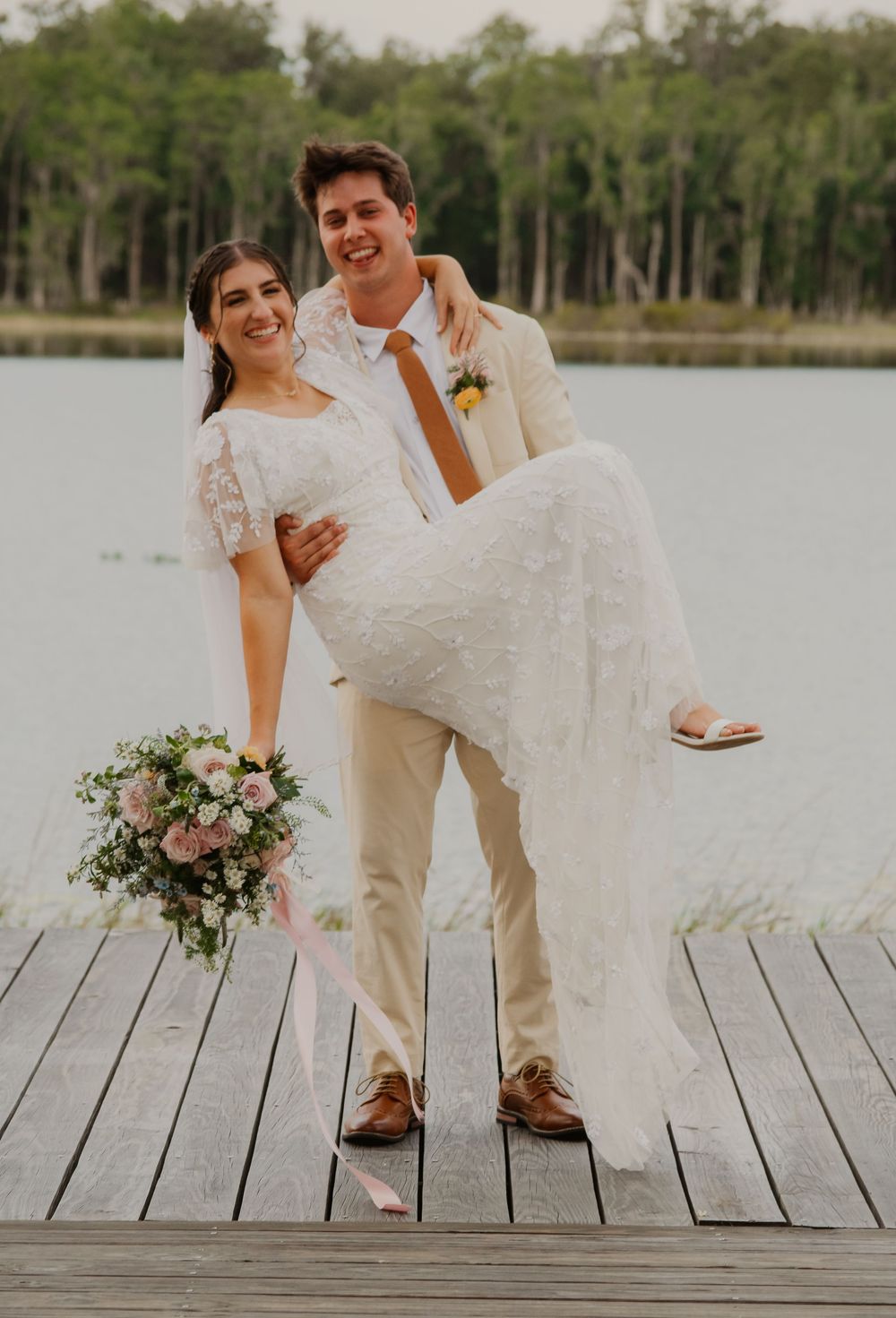 Smiling groom in tan suit carries bride in white lace dress on a wooden dock with a serene lake in the background.