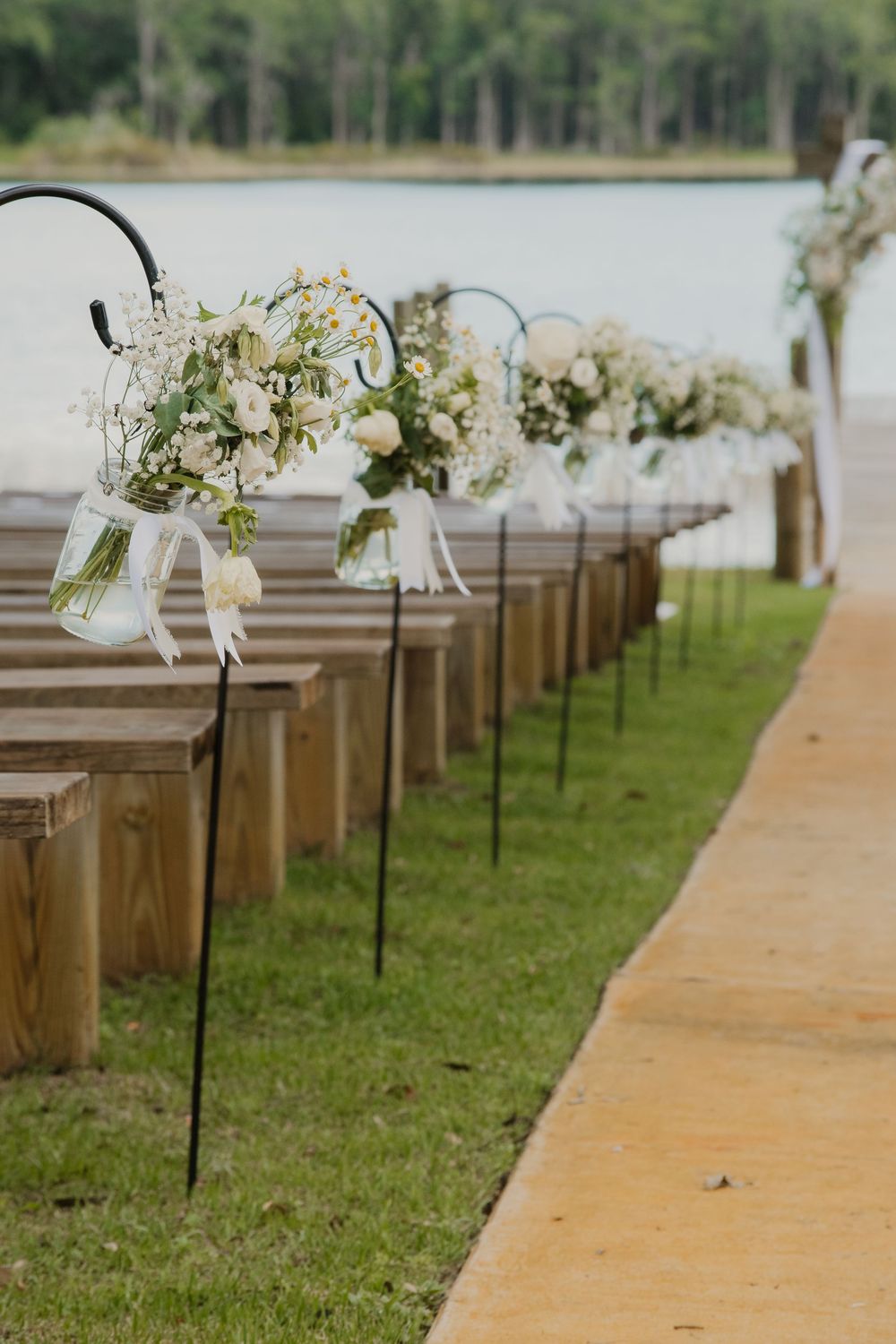 Elegant outdoor wedding ceremony aisle lined with wooden pews and white floral arrangements on shepherd hooks near a lake.