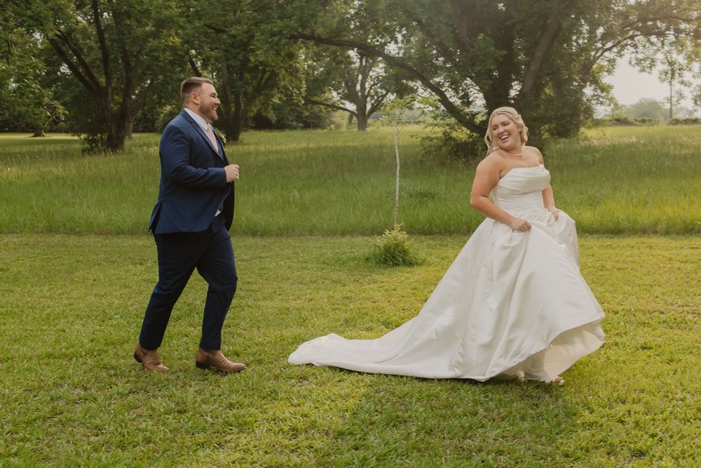 Joyful bride lifting her white ballgown while groom approaches smiling during first look in a green meadow.