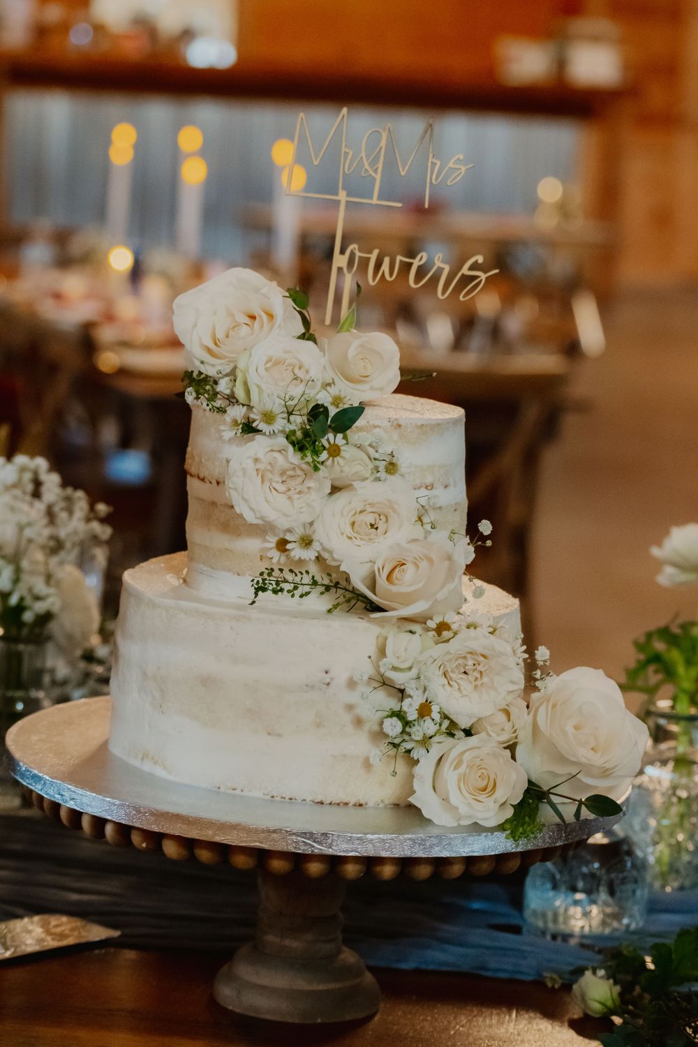 Two-tier semi-naked wedding cake adorned with white roses and greenery, topped with gold Mr and Mrs cake topper.