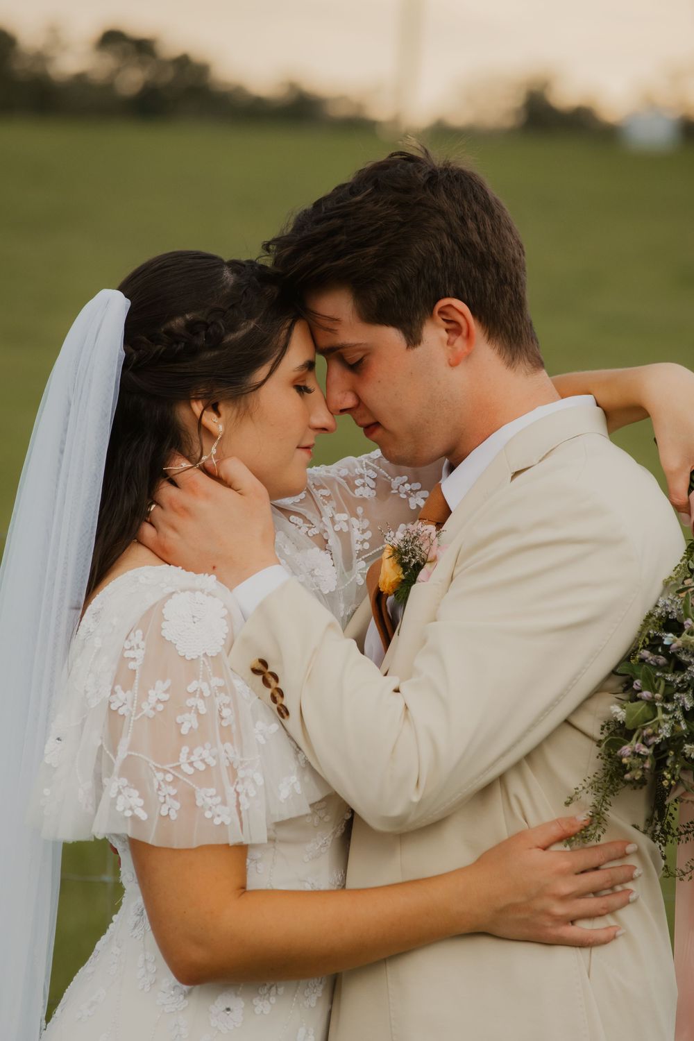 Bride in lace veil and groom in ivory suit share an intimate forehead-to-forehead moment in a green field at sunset.
