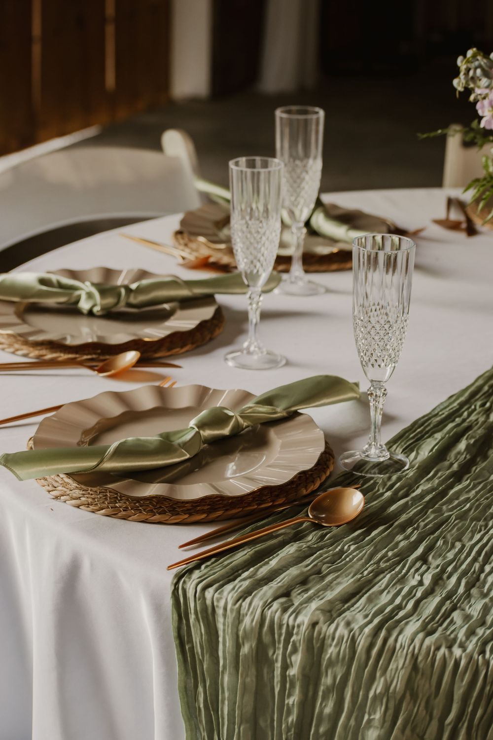 Elegant wedding table setting with sage green napkins, rattan chargers, crystal champagne flutes, and gold cutlery.