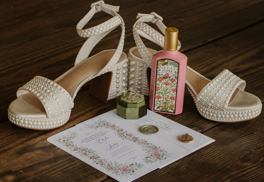 Pearl-embellished bridal heels with ankle straps beside a Gucci perfume bottle, ring box, and floral wedding invitation on wood.