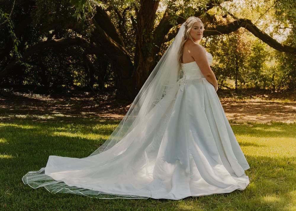 Elegant bride in a strapless light blue ballgown with cathedral veil posing in a lush green garden setting.