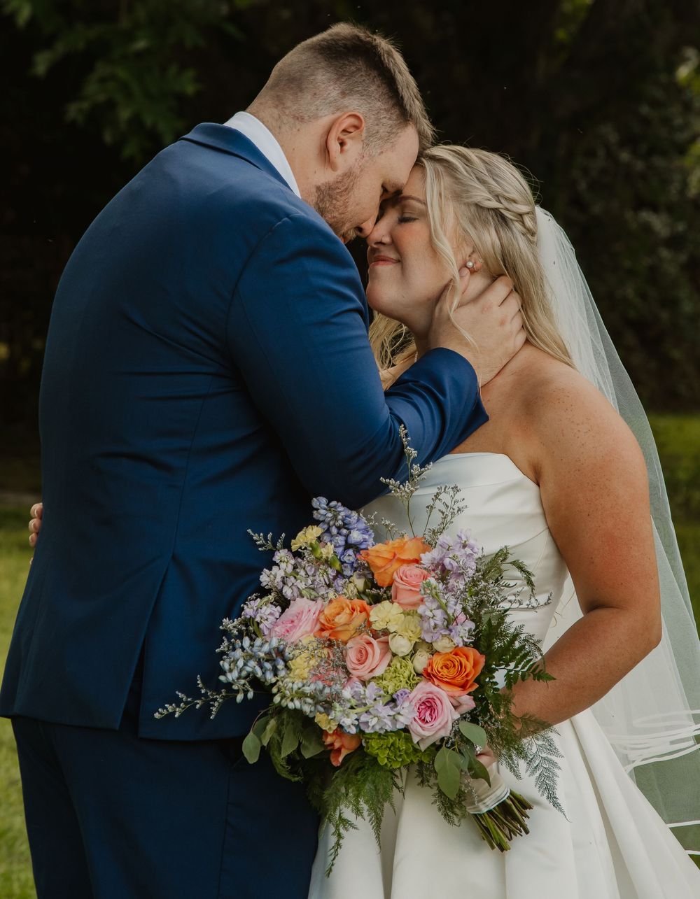 Bride and groom sharing a tender forehead-to-forehead moment, bride holding a colorful spring wedding bouquet outdoors.