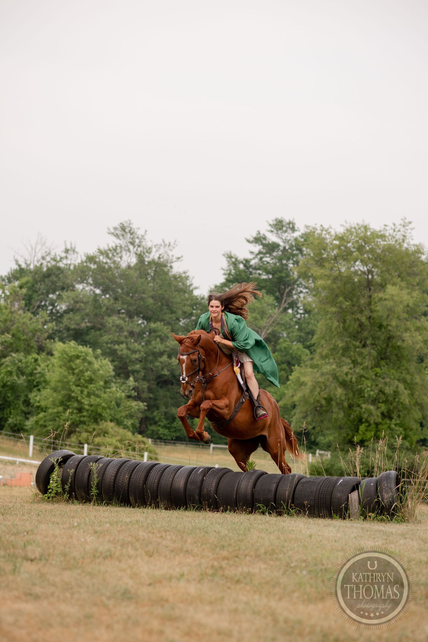 A chestnut horse and rider jumping over lined up tires in a grassy field.
