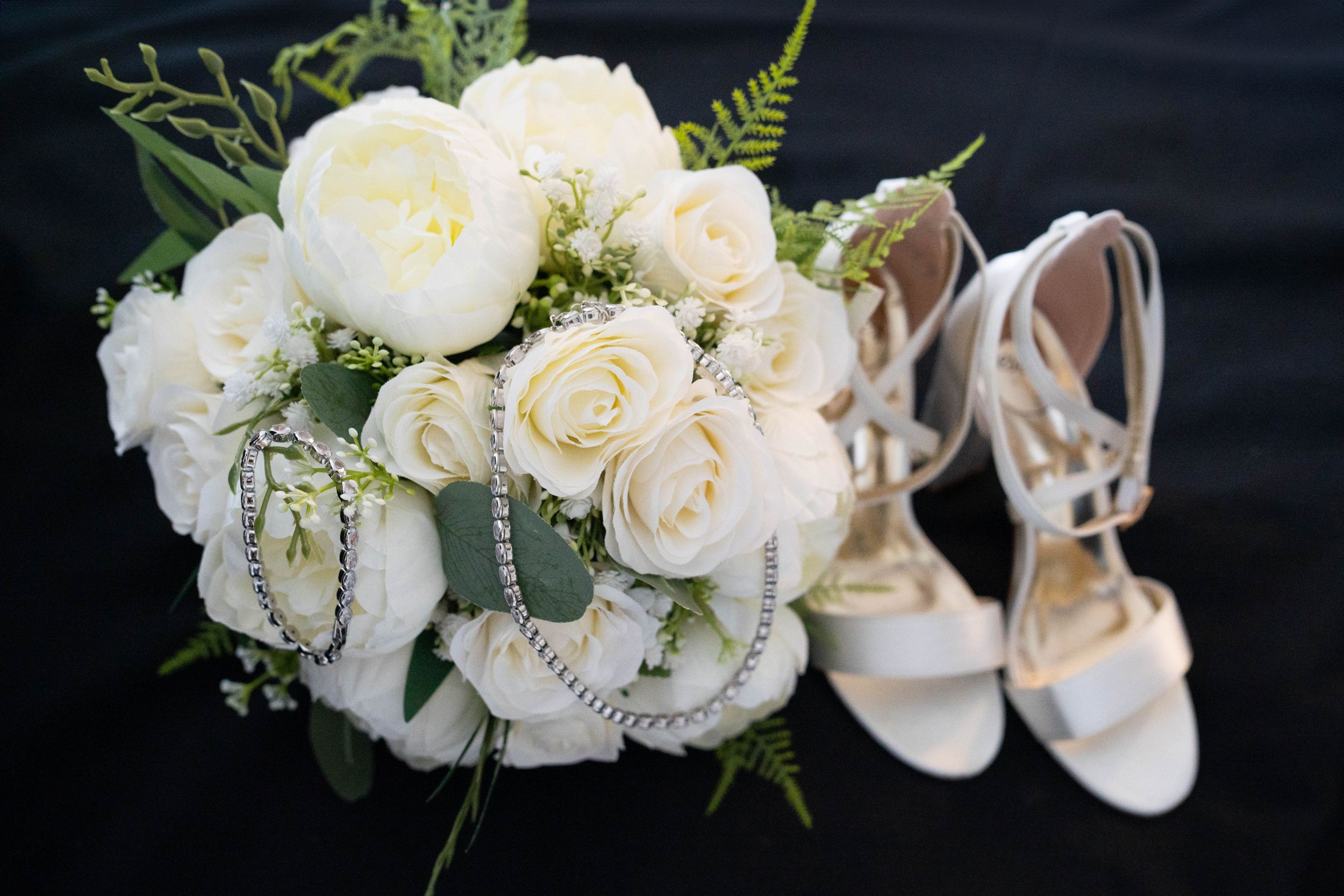 White rose bridal bouquet displayed next to elegant strappy white wedding heels on black background.