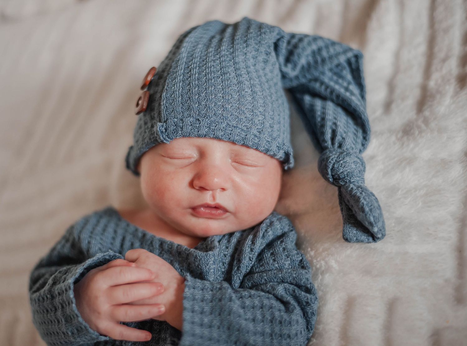 Sleeping baby in blue knitted outfit and matching cap on soft bedding.