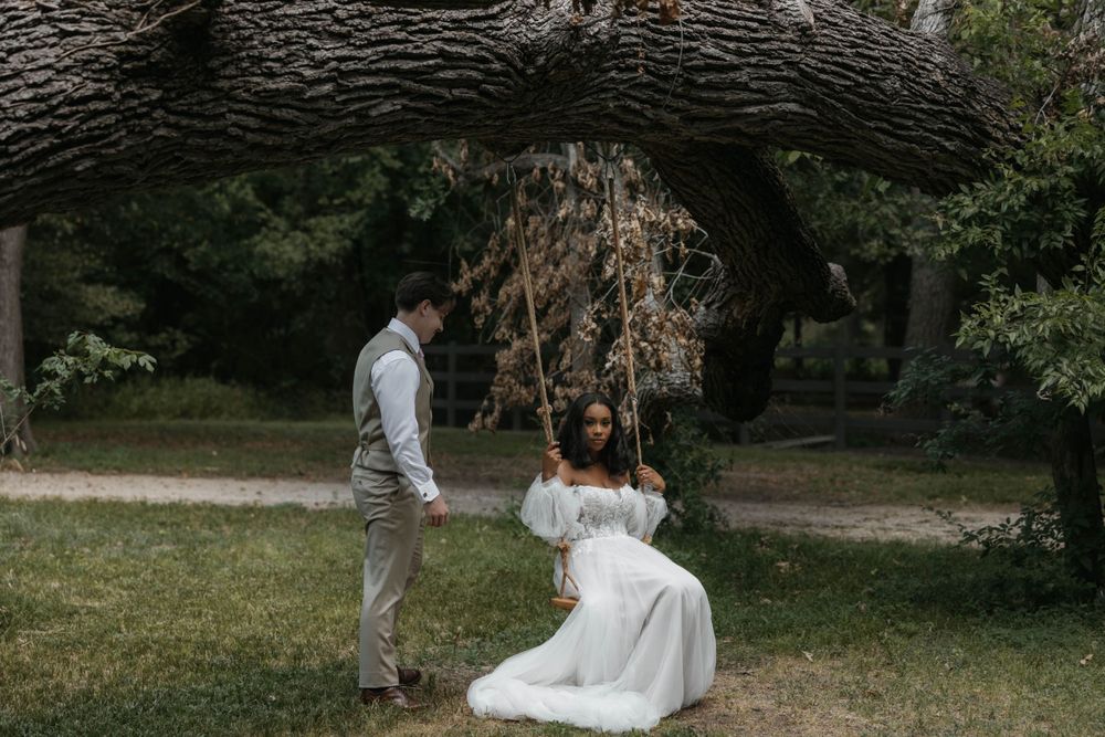 Bride in white gown sitting on tree swing while groom stands nearby in lush garden.