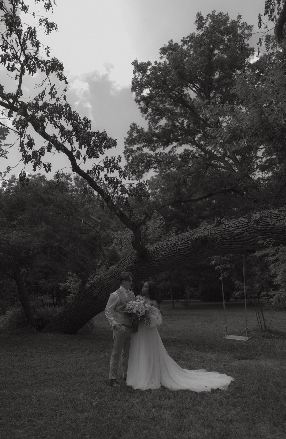 Bride and groom embracing under a large fallen tree in a park, black and white photo.