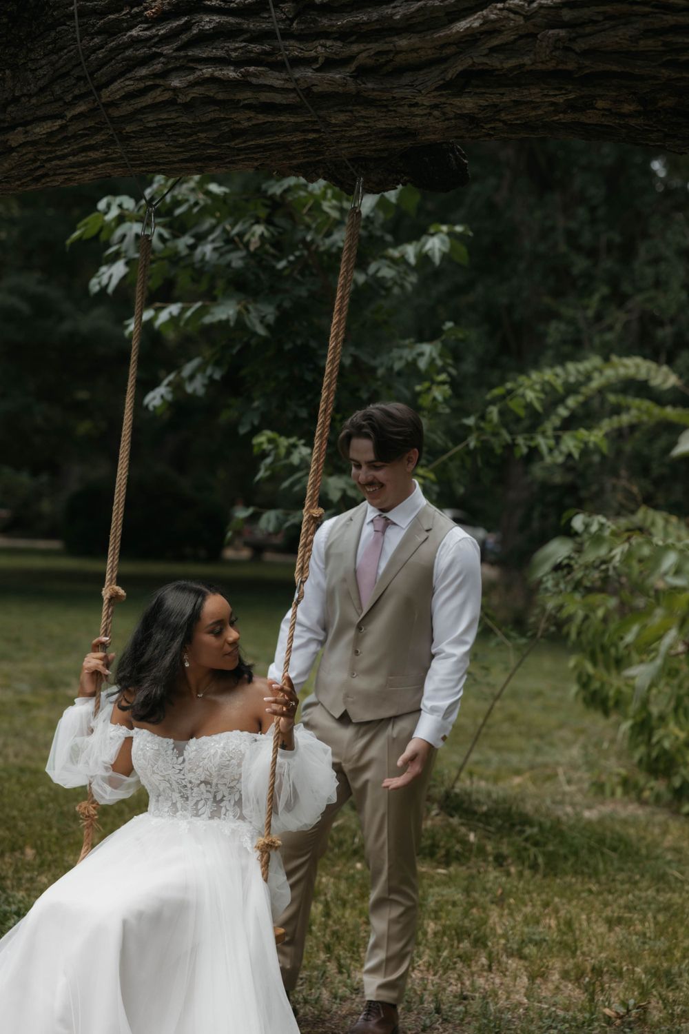 Bride in lace gown sitting on rope swing under tree as groom smiles beside her.
