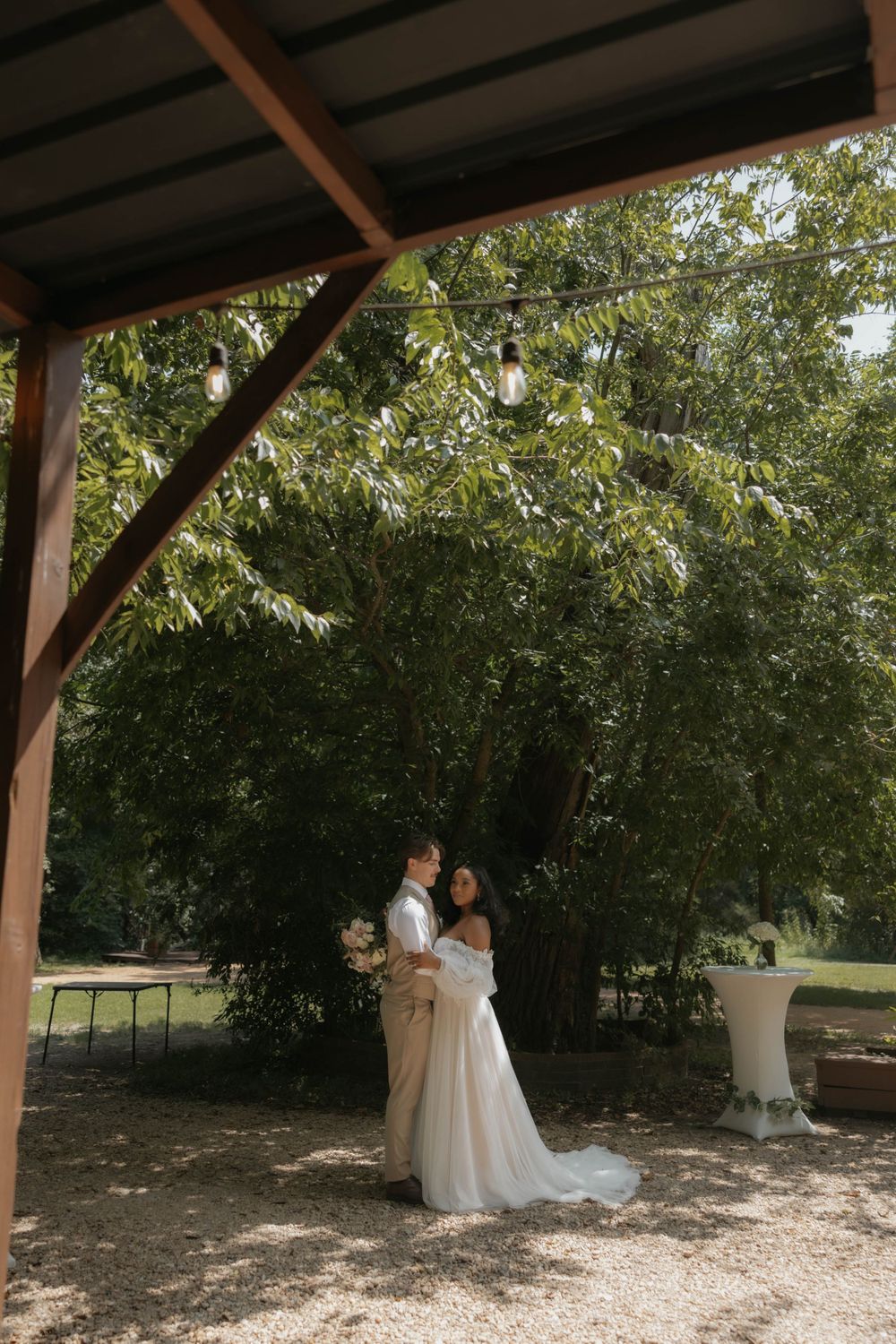 Bride and groom embracing under string lights on a wooden pergola in a garden venue.