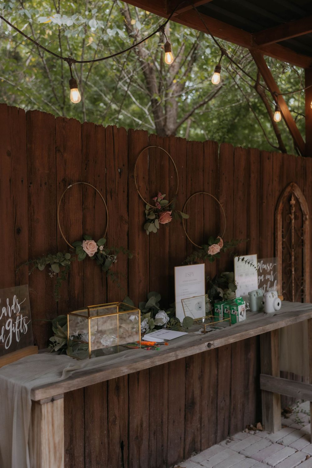 Rustic outdoor wedding gift table with gold hoop floral decor and Edison bulb string lights.
