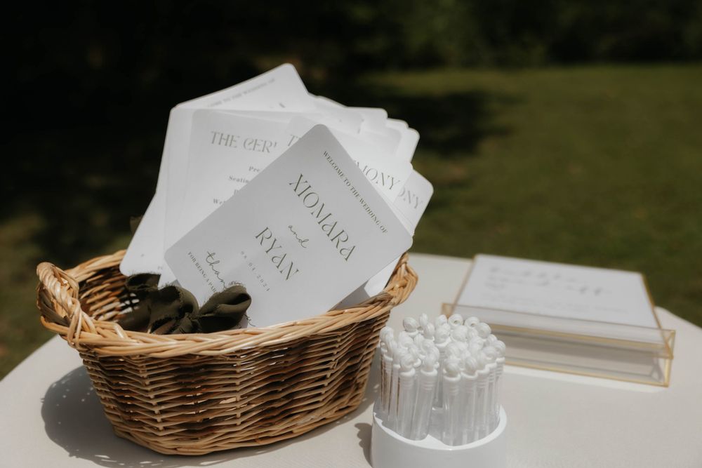 Wedding ceremony programs in a wicker basket with couple names Nomara and Ryan on white cards.