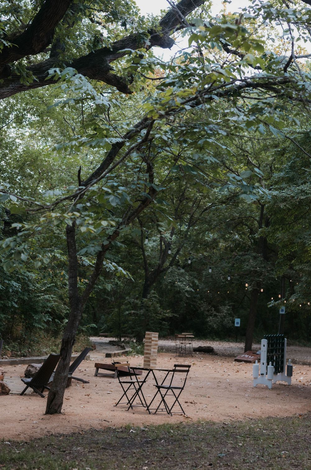 Outdoor wedding lounge area with bistro chairs, giant Jenga, and string lights under forest trees.