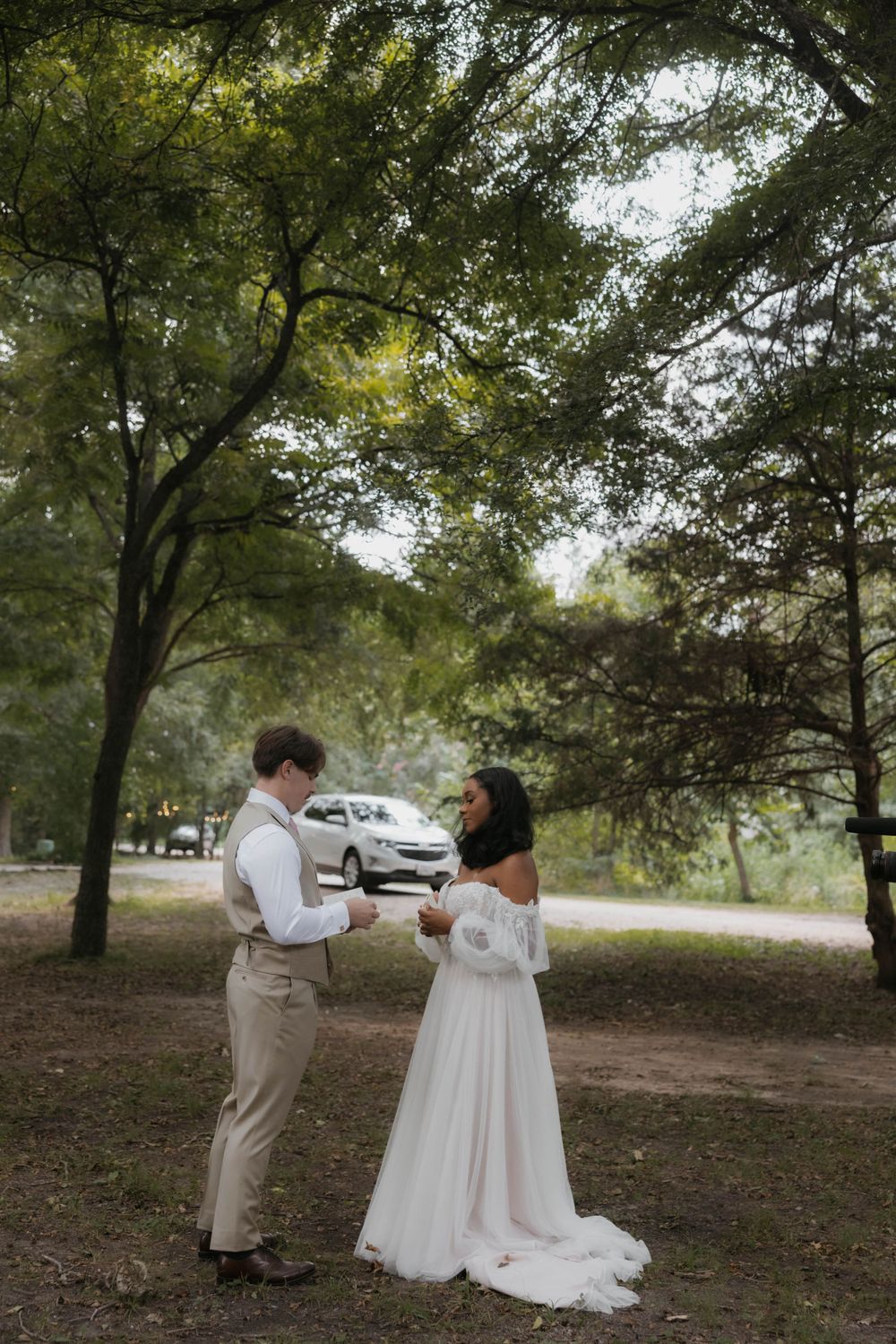 Bride and groom share a first look moment beneath lush green trees in outdoor wooded setting.