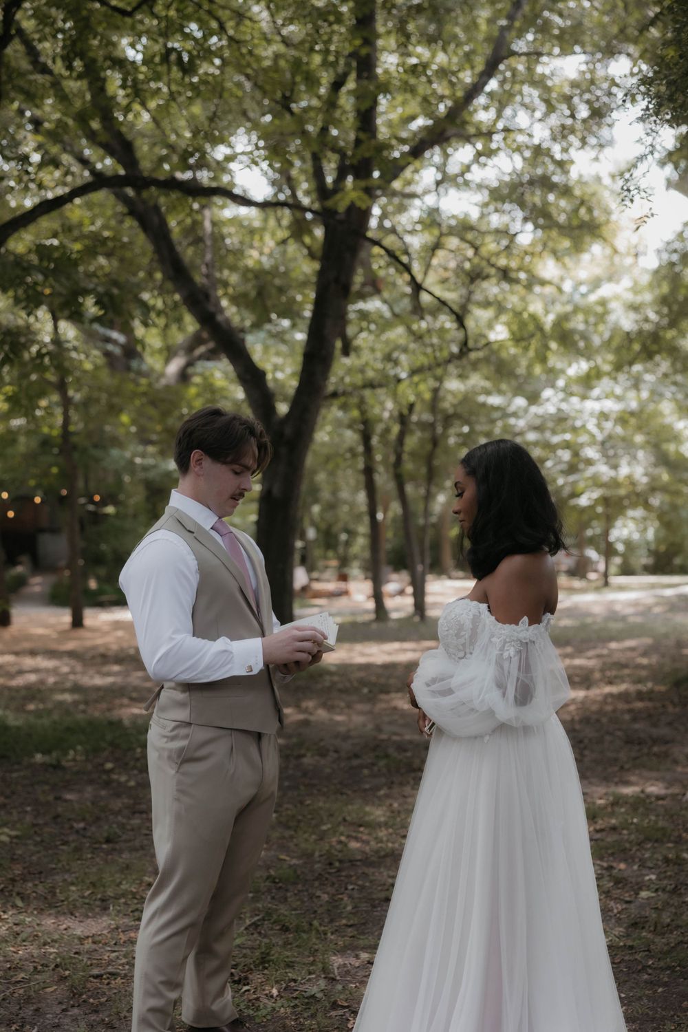 Bride in off-shoulder lace gown and groom in tan suit exchange vows outdoors in a wooded garden setting.