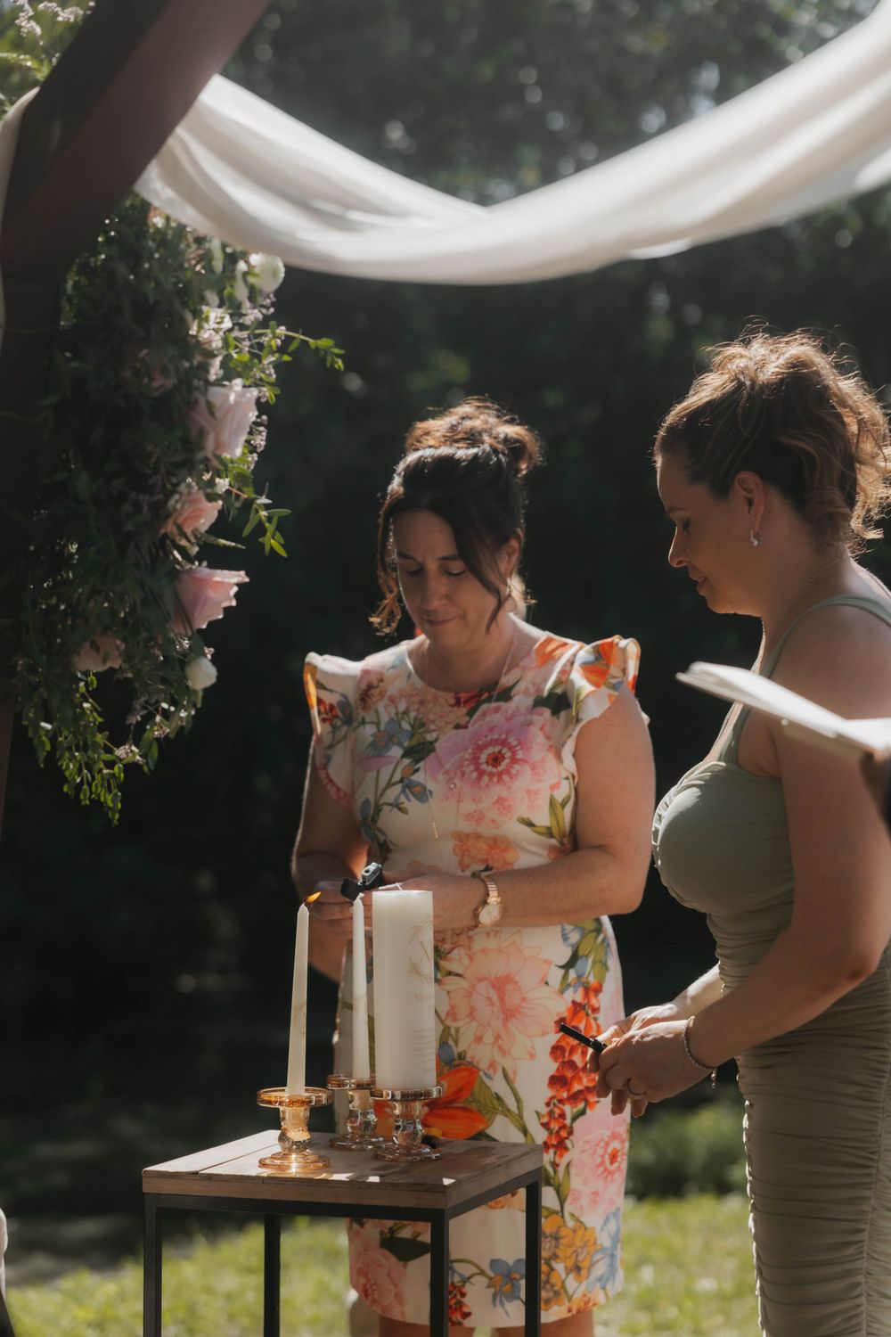 Two women lighting white pillar candles at an outdoor wedding ceremony beneath a floral arch.