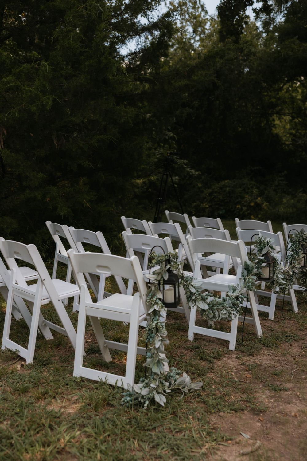 White folding chairs with greenery garlands and lanterns set up for outdoor forest wedding ceremony.
