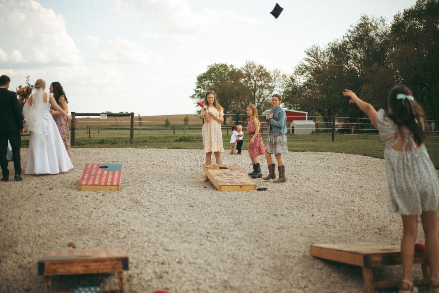Wedding guests playing outdoor games with cornhole boards on a gravel surface.