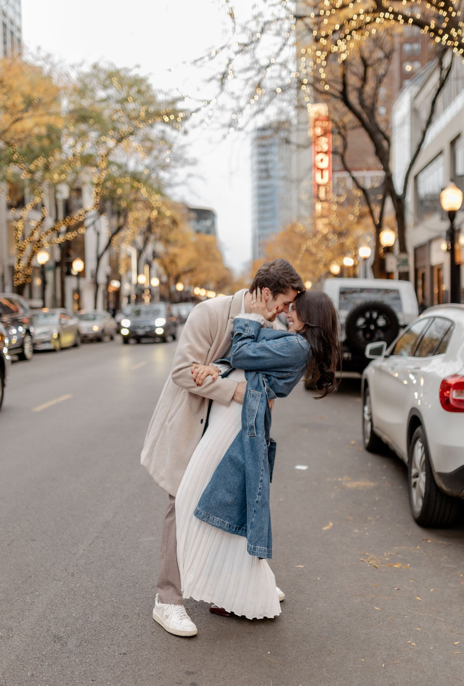 A romantic embrace on an urban street lined with autumn trees and parked cars.