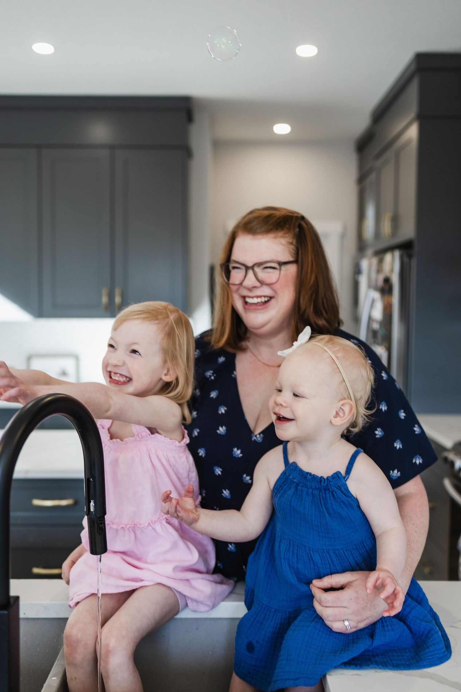 A mother and her two daighters delight in bubbles being blown toward them in the kitchen