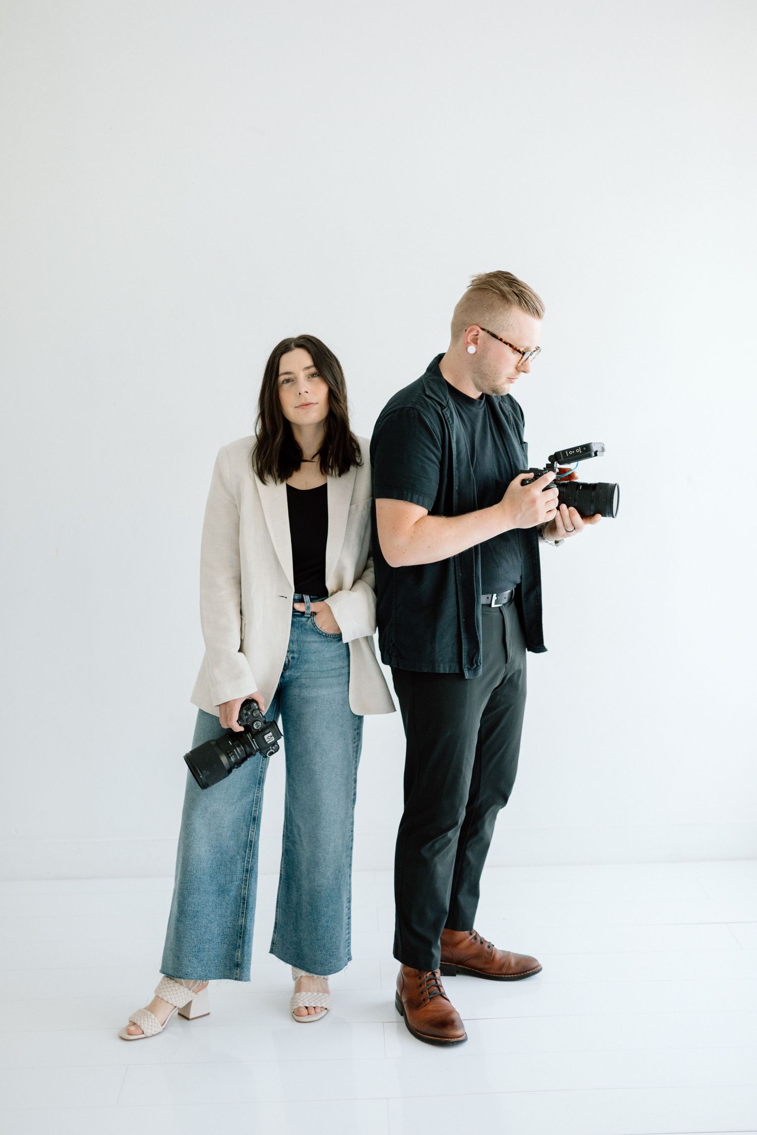Two photographers with cameras standing against white backdrop in casual attire.