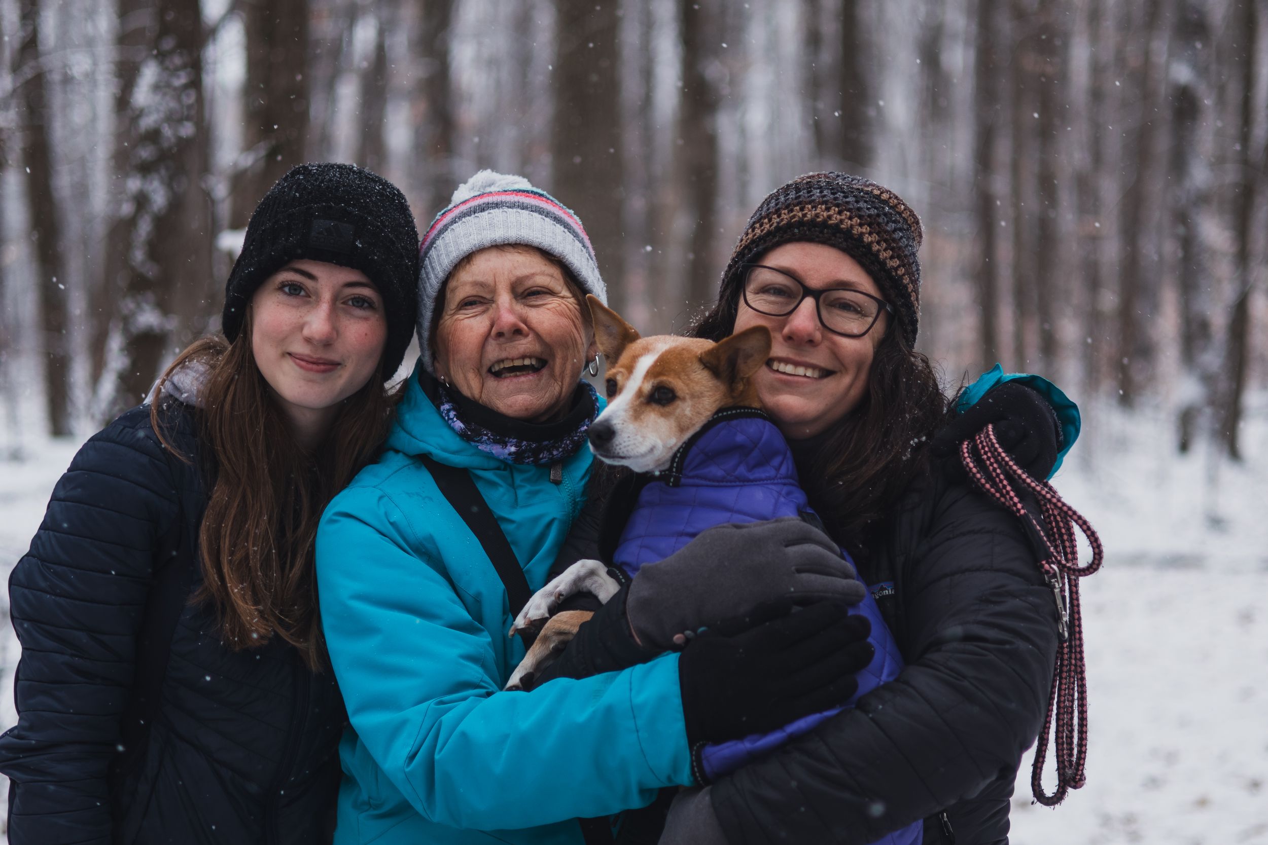 A group of people bundled in winter clothing smile together in a snowy forest setting.