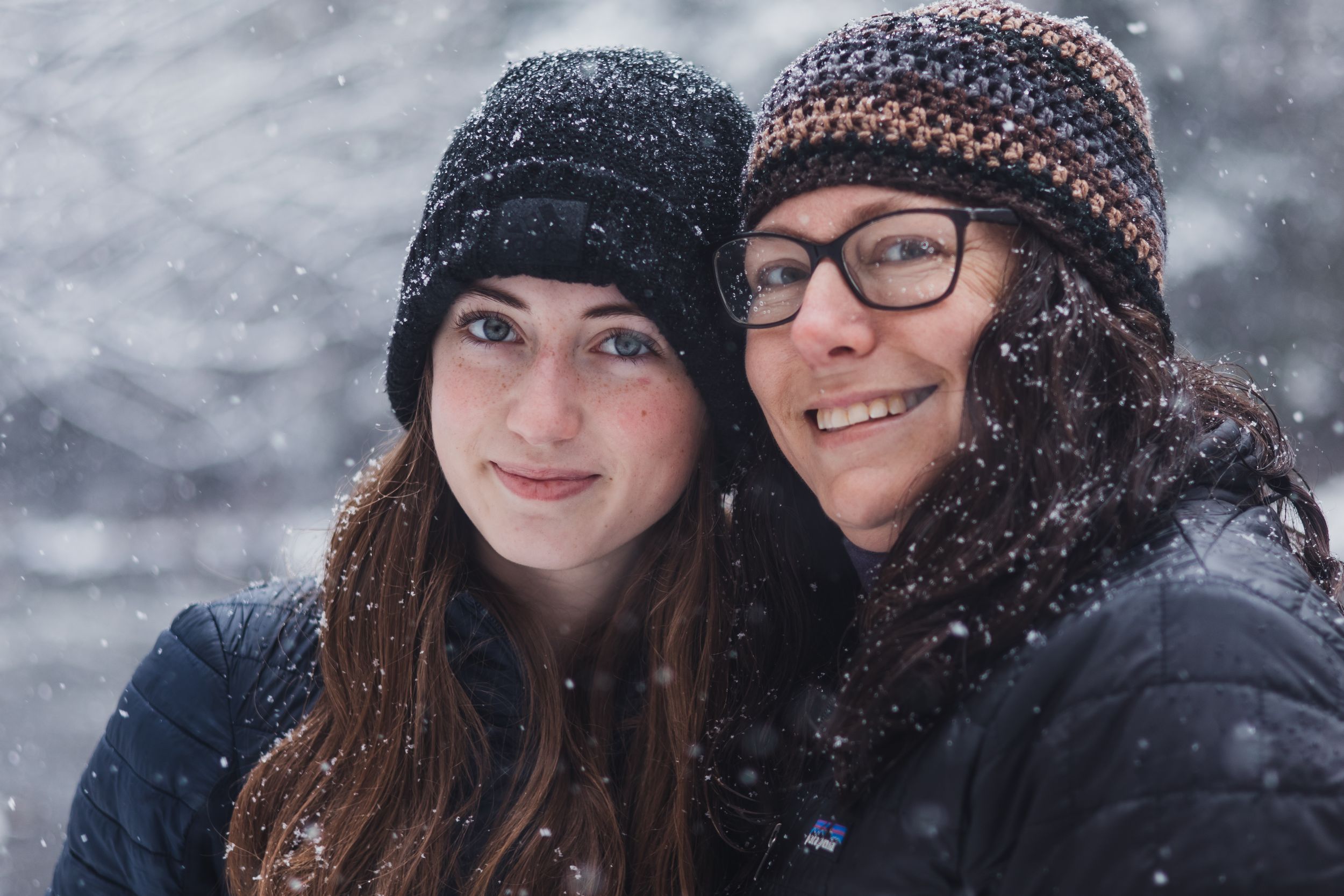 Two people wearing winter hats smile together while snow falls around them in a wintry outdoor scene.