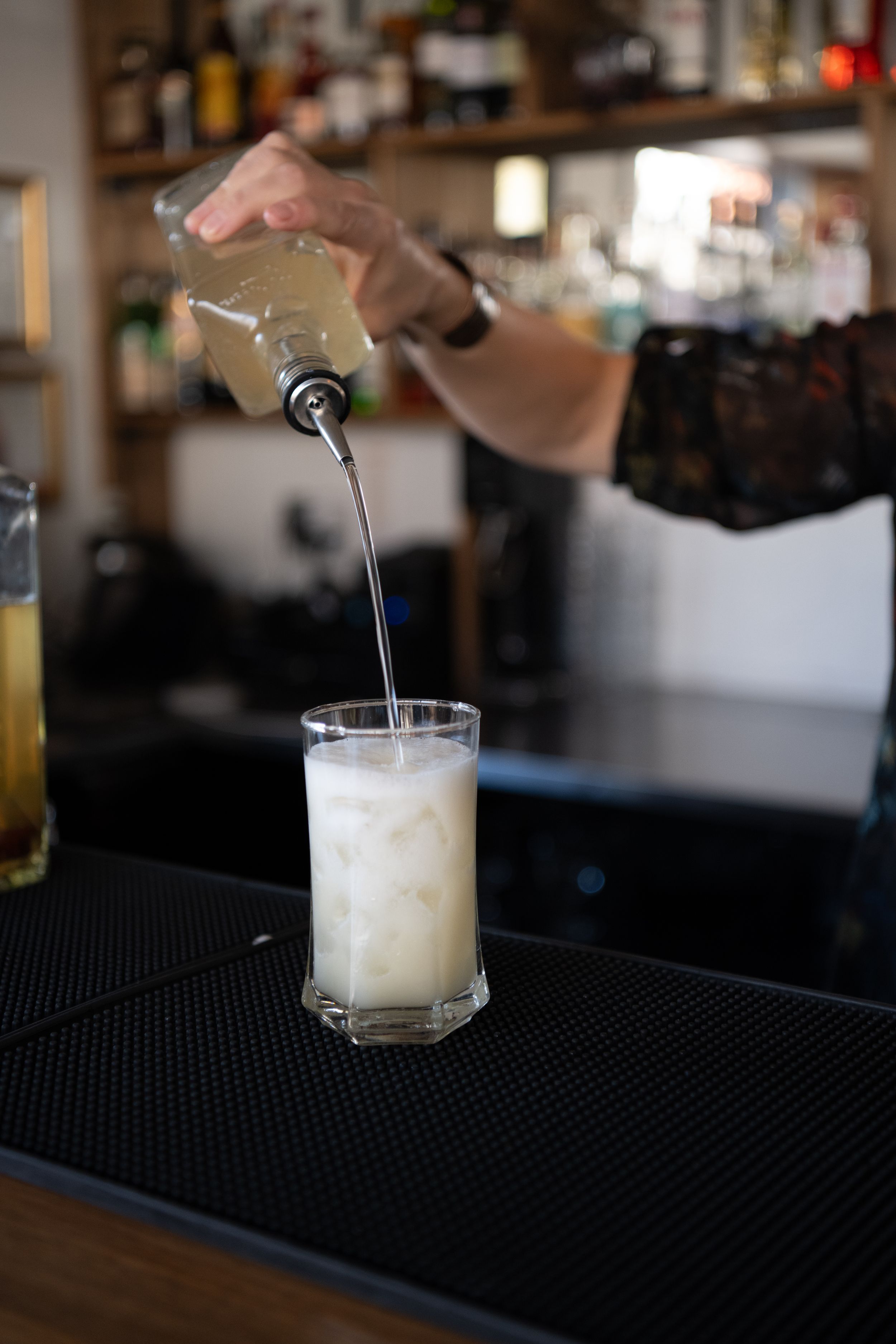 Bartender pouring a creamy white cocktail into a glass at a dimly lit bar counter.