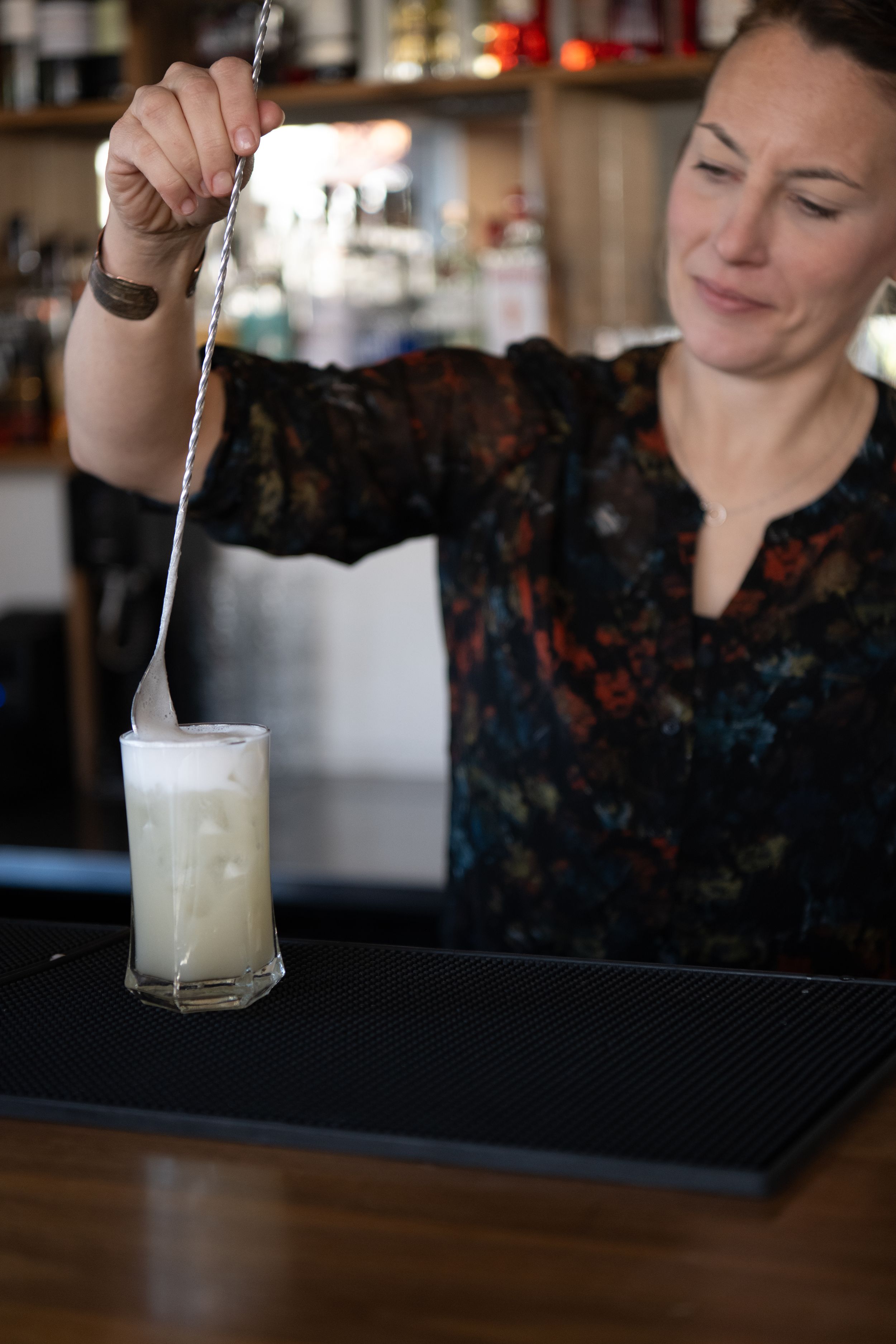 A bartender pours a frothy white cocktail into a glass at a dimly lit bar counter.