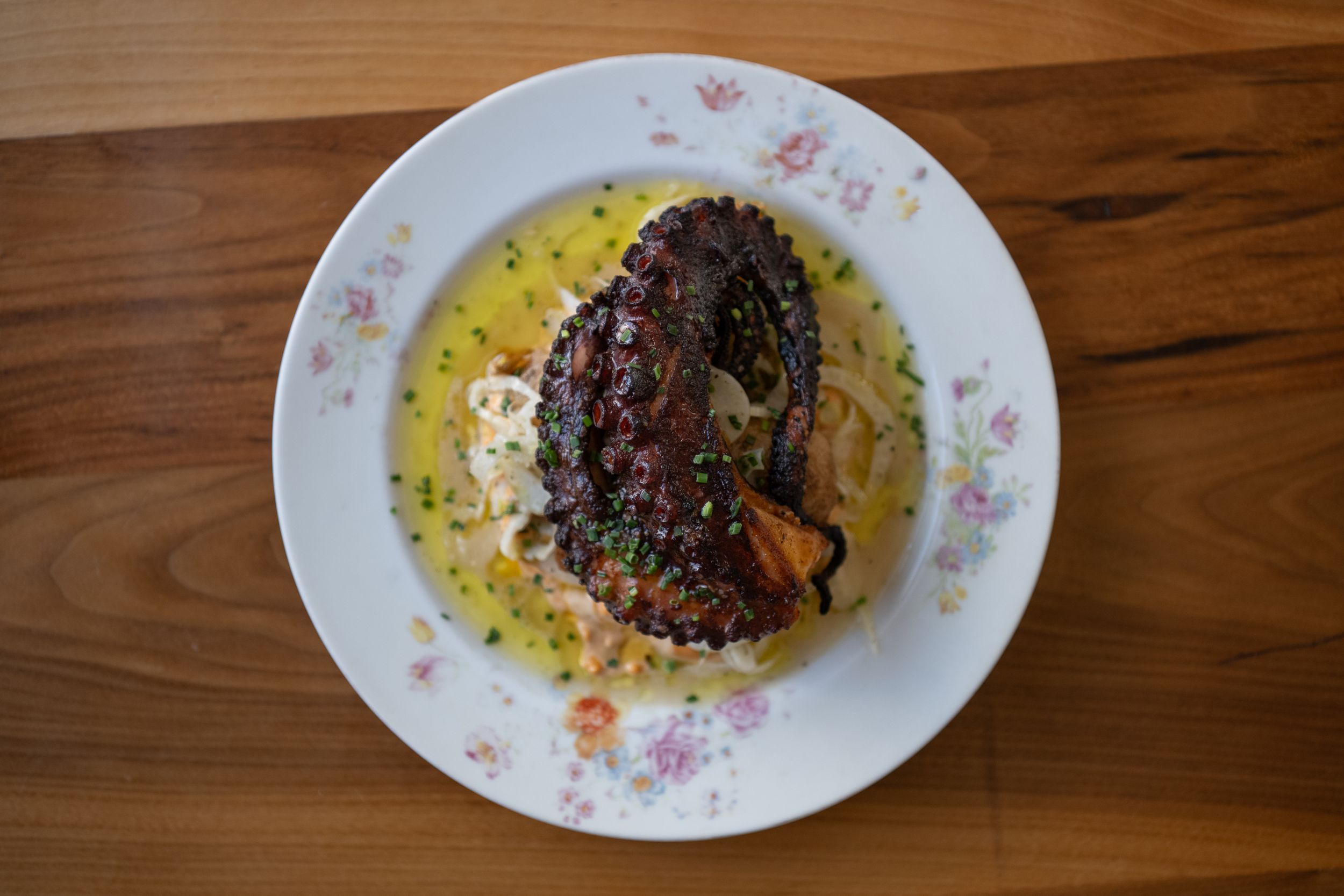Plated dish with dark garnish on floral-patterned oval plate against wooden surface with yellow sauce.