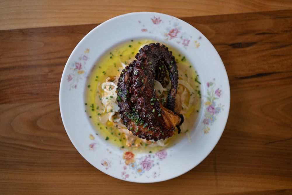 Plated dish with dark garnish on floral-patterned oval plate against wooden surface with yellow sauce.