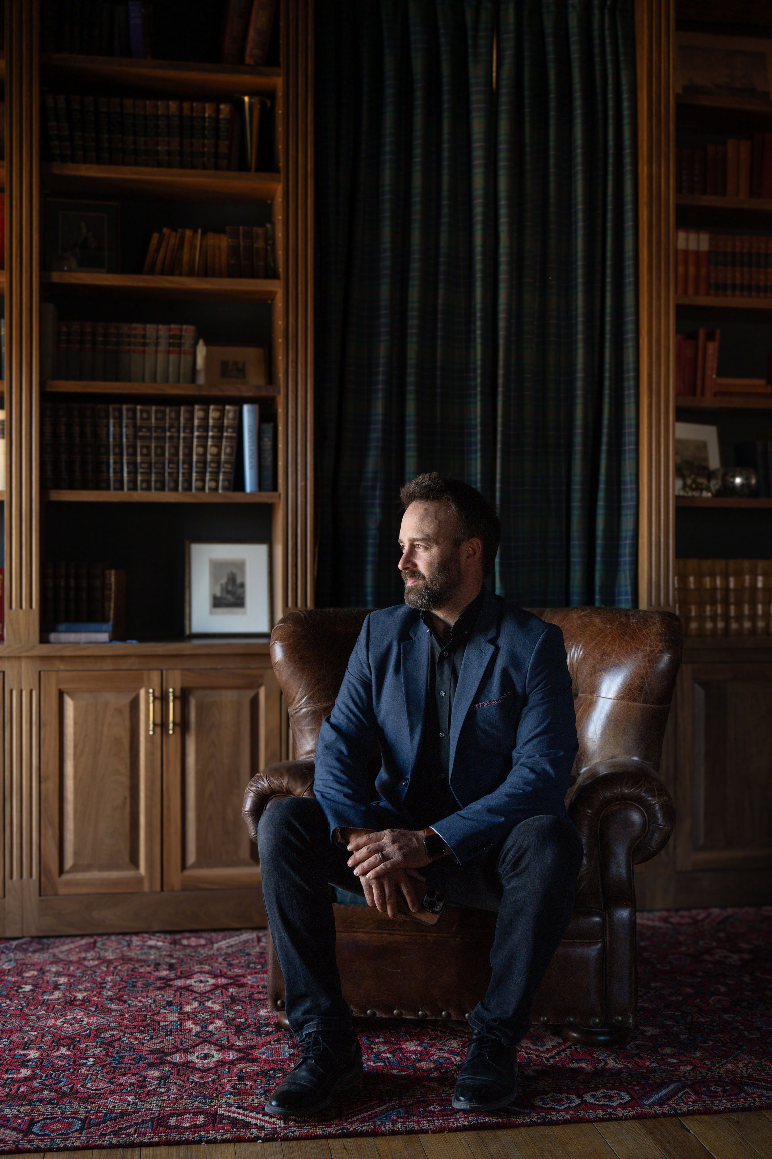 A person in a dark suit sits contemplatively on a leather chair in an elegant library with wood paneling and bookshelves.