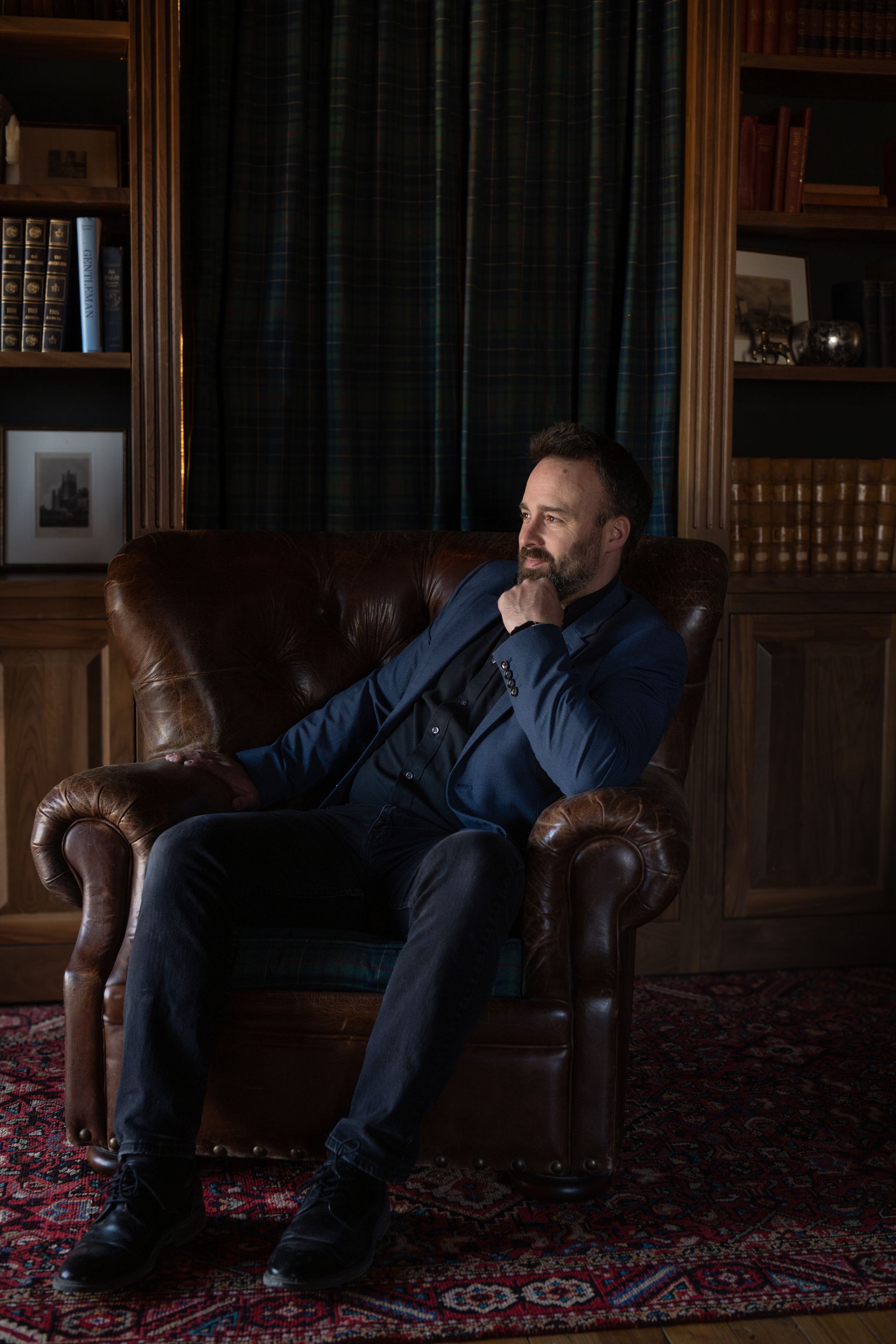 A person in a dark suit reclines on a leather chair in an elegant library with a patterned rug.