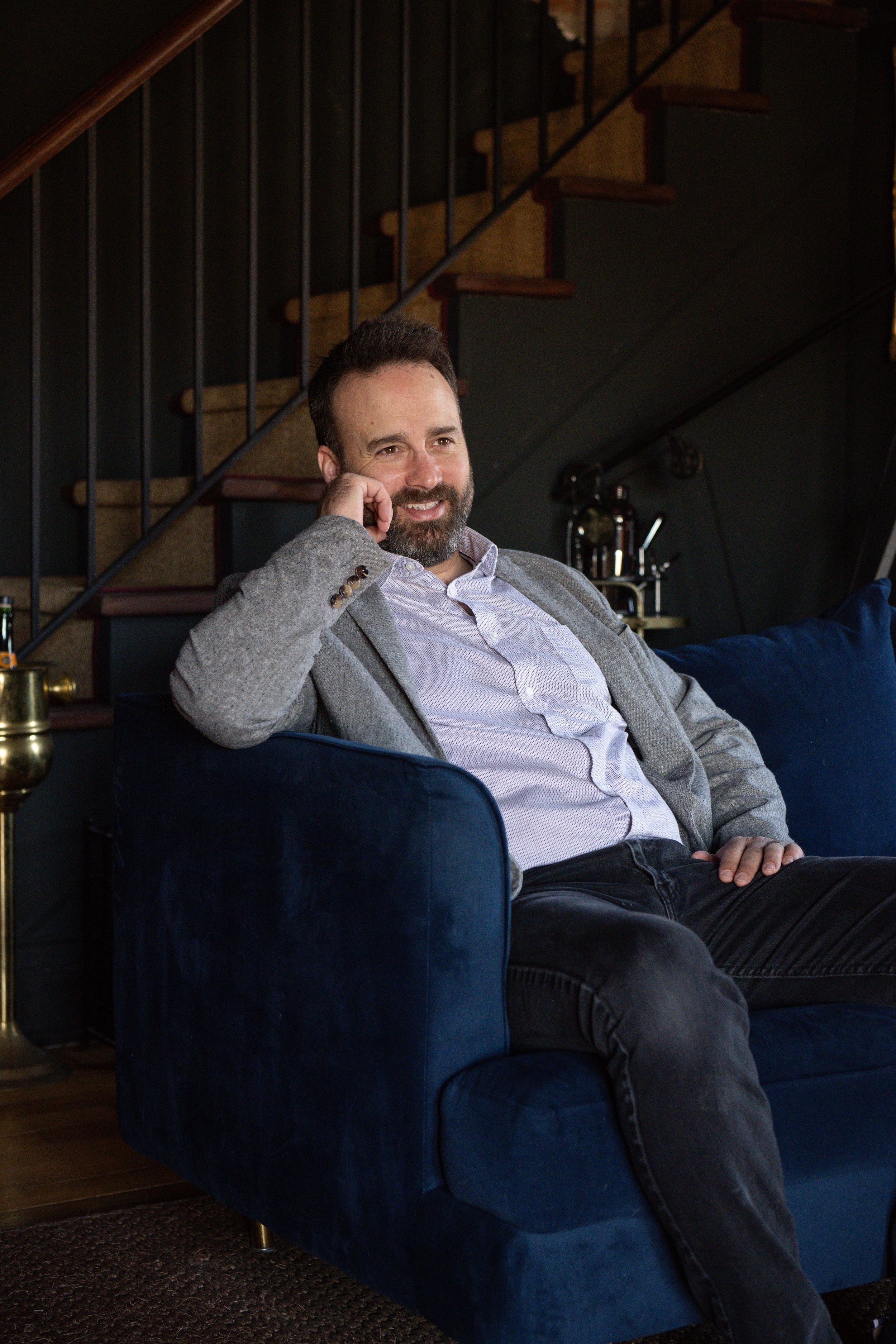 A person relaxes on a navy blue couch while laughing and sitting against wooden stairs in a dimly lit room.