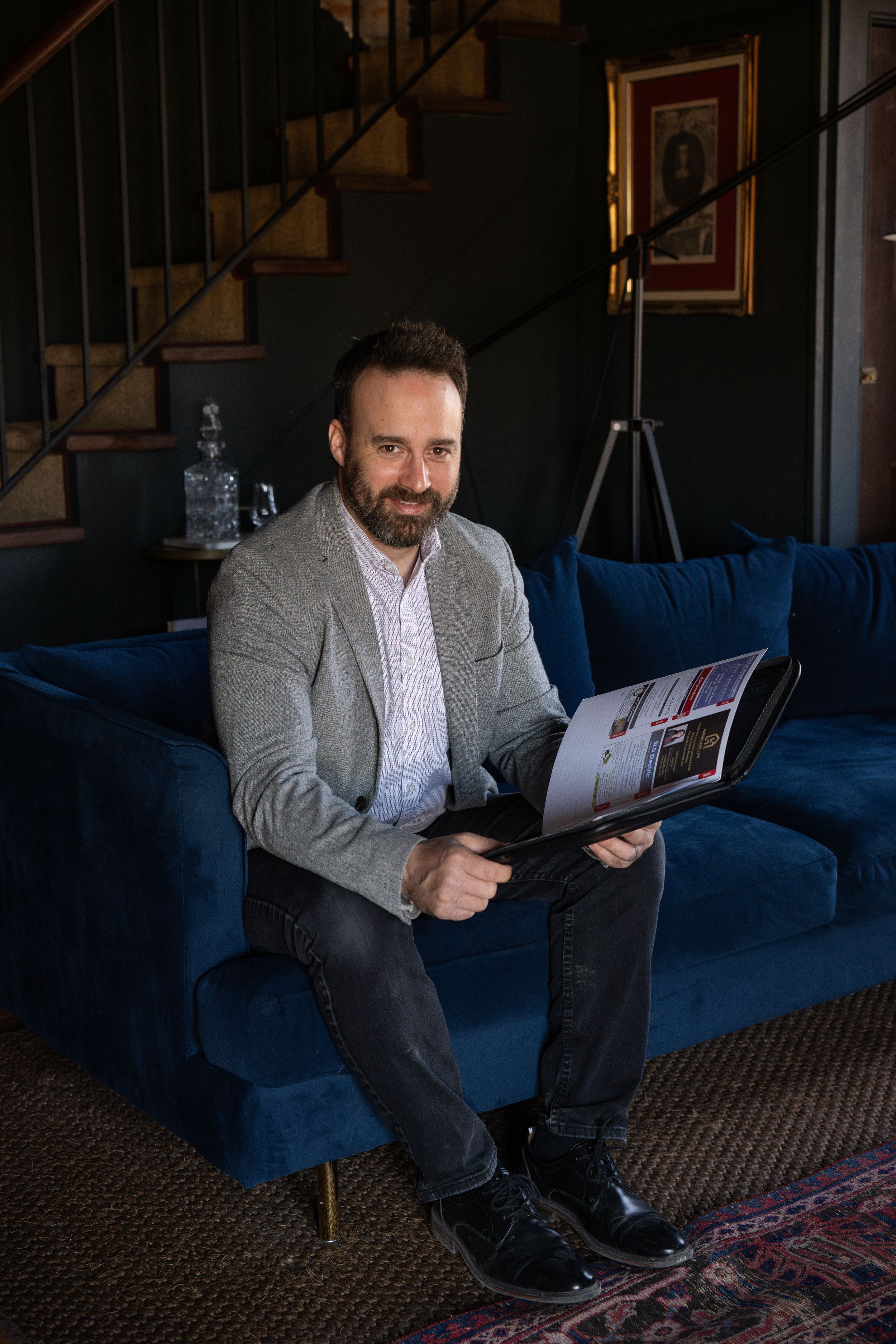 A person sits on a blue velvet couch holding a magazine in a dark room with stairs and framed art visible. Quinte West