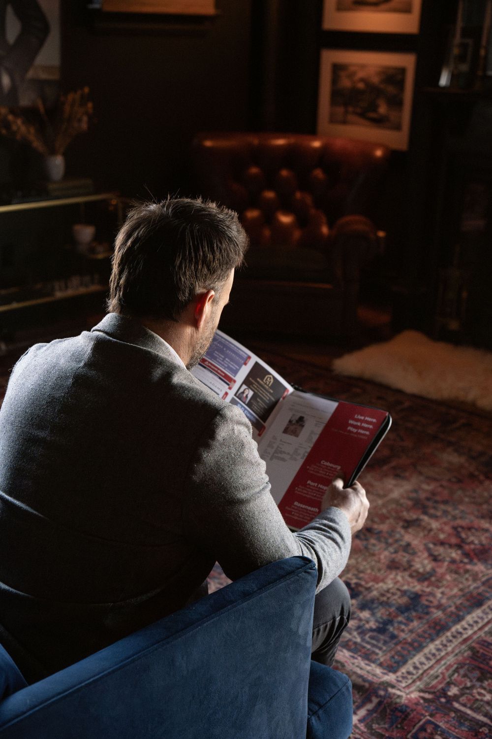 Someone reads a book while relaxing in a cozy armchair on a patterned rug in a dimly lit room with framed artwork.